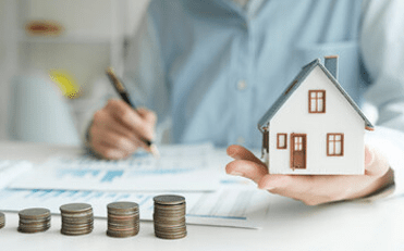 A man holding a model house while writing on a spreadsheet with progressively taller stacks of coins on a table, symbolizing the financial benefits of energy-efficient home upgrades.