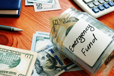 A jar of emergency savings money tipped over on a table with scattered bills, a calculator, checkbook, and pen.